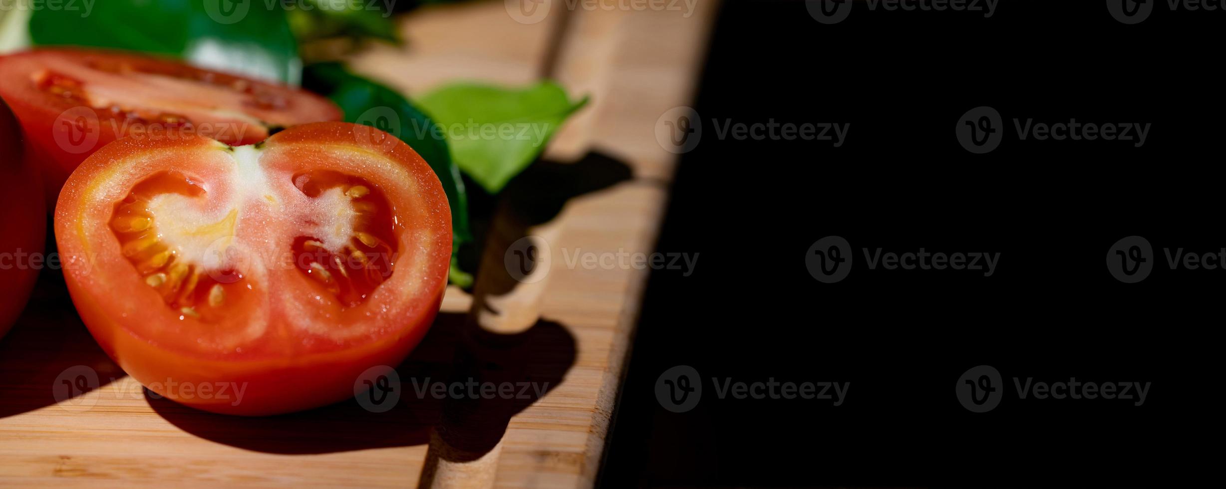 tomate e metade do tomate fatiado ao lado, na placa de madeira na luz do estúdio com tema escuro. foto