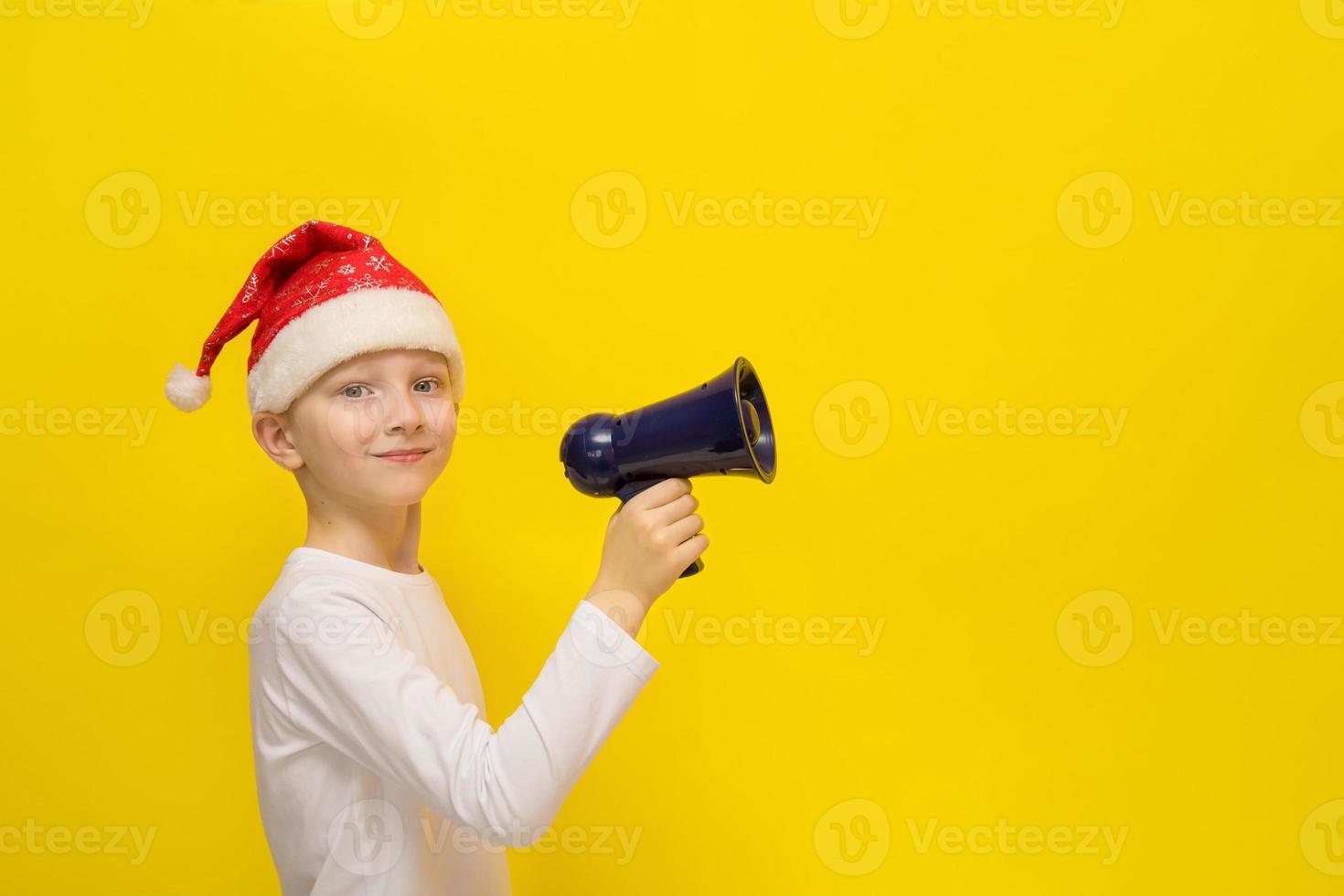 menino com um chapéu de papai noel segura um megafone em suas mãos em um fundo amarelo com espaço de cópia, natal, férias de inverno foto