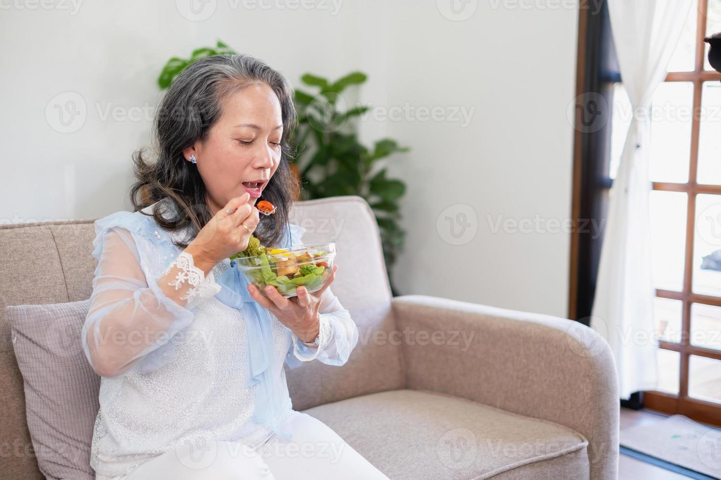 mulher asiática sênior sentada comendo salada de legumes e comida saudável e comendo alegremente no sofá da casa para um corpo saudável. conceito de comida saudável foto