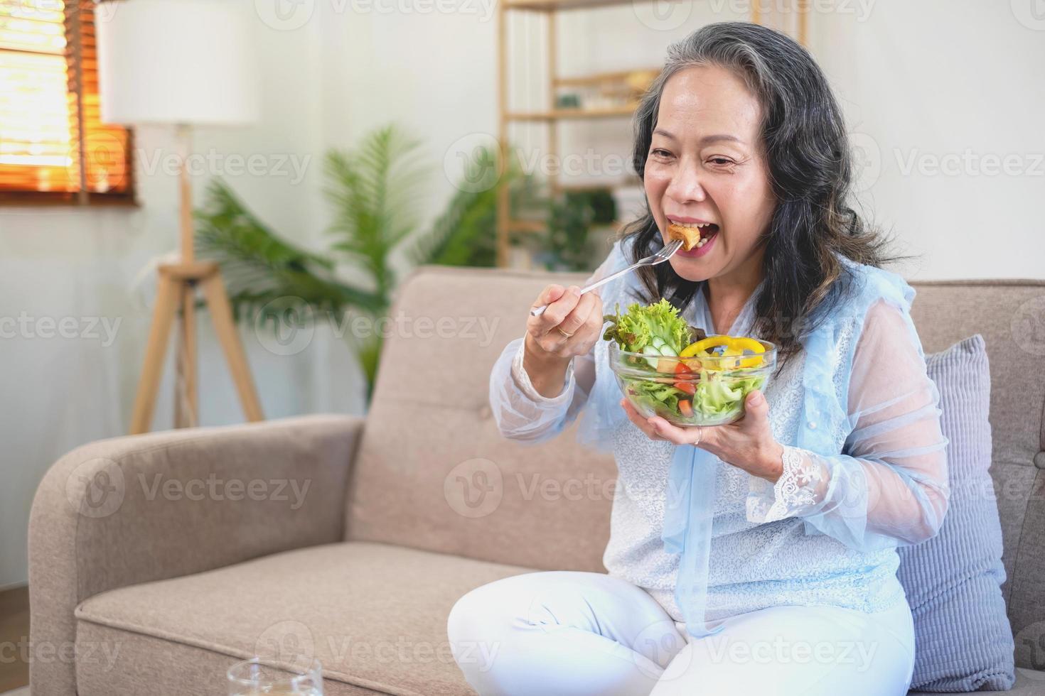 mulher asiática sênior sentada comendo salada de legumes e comida saudável e comendo alegremente no sofá da casa para um corpo saudável. conceito de comida saudável foto