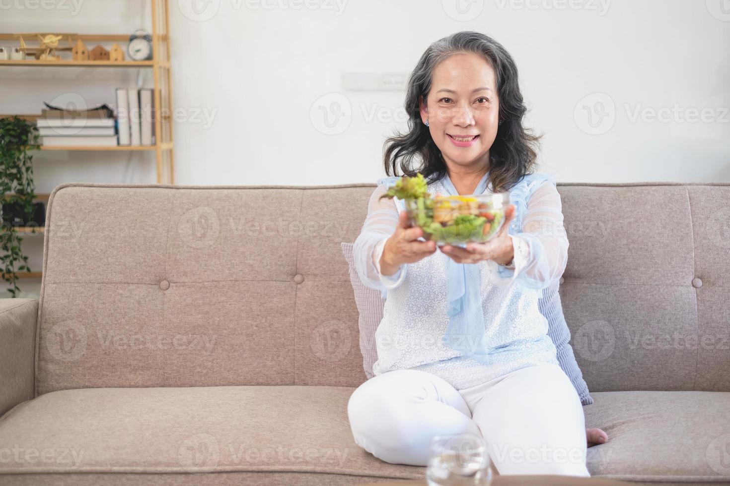 mulher asiática sênior sentada comendo salada de legumes e comida saudável e comendo alegremente no sofá da casa para um corpo saudável. conceito de comida saudável foto