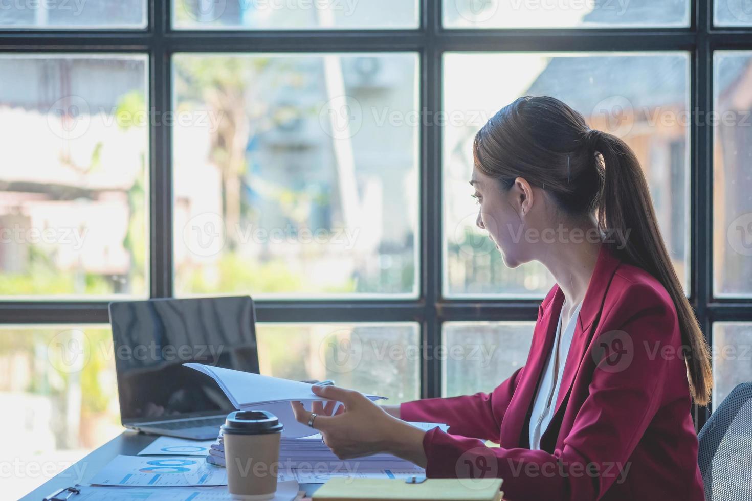 financeira, empresária asiática de terno vermelho segurando a xícara de café sentada na mesa no escritório, tendo computador para fazer trabalho de contabilidade no local de trabalho para calcular o lucro anual por dever, ideia de negócio foto