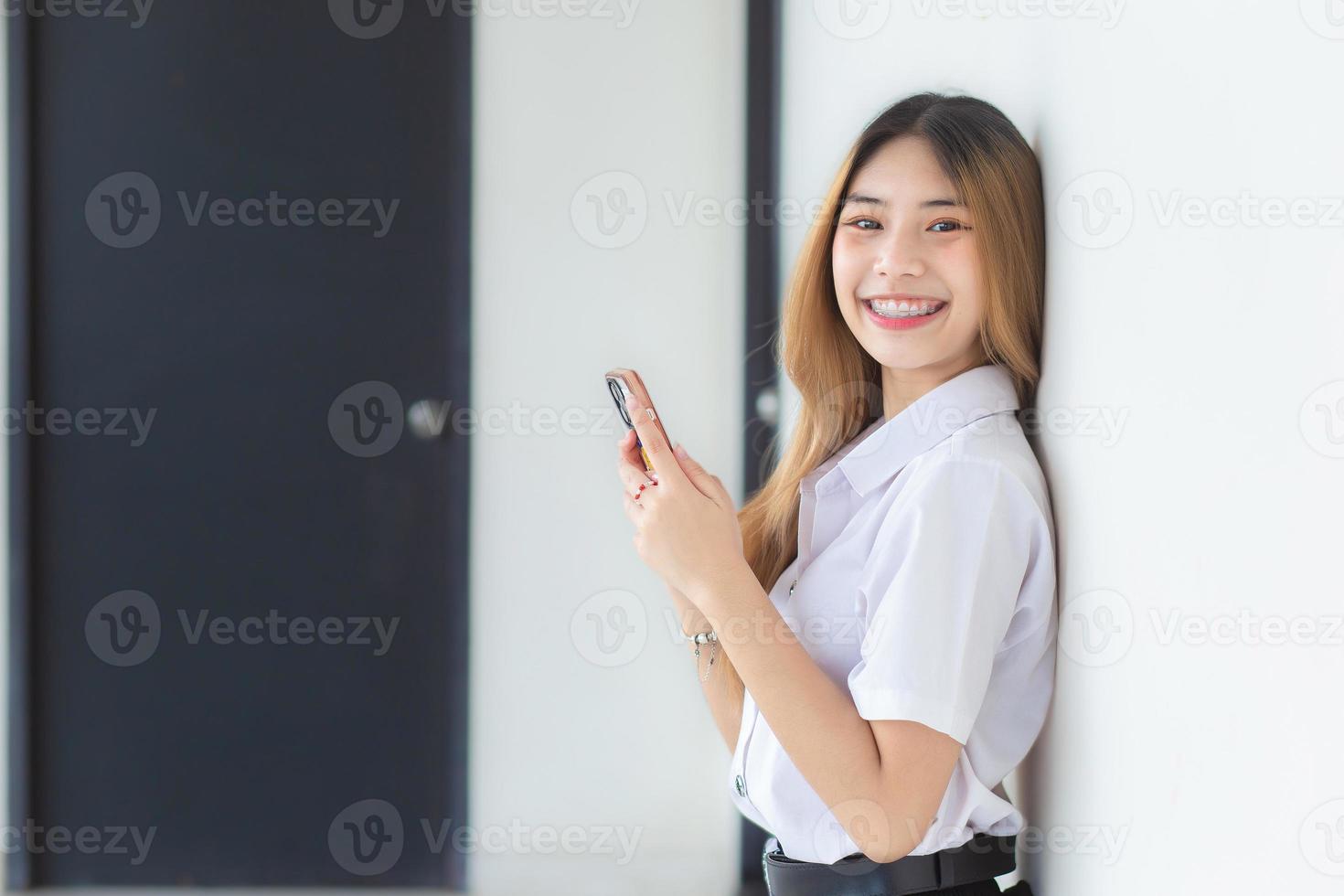 o retrato da jovem estudante tailandesa asiática linda e muito fofa de uniforme está sorrindo alegremente enquanto usa um smartphone na universidade. foto