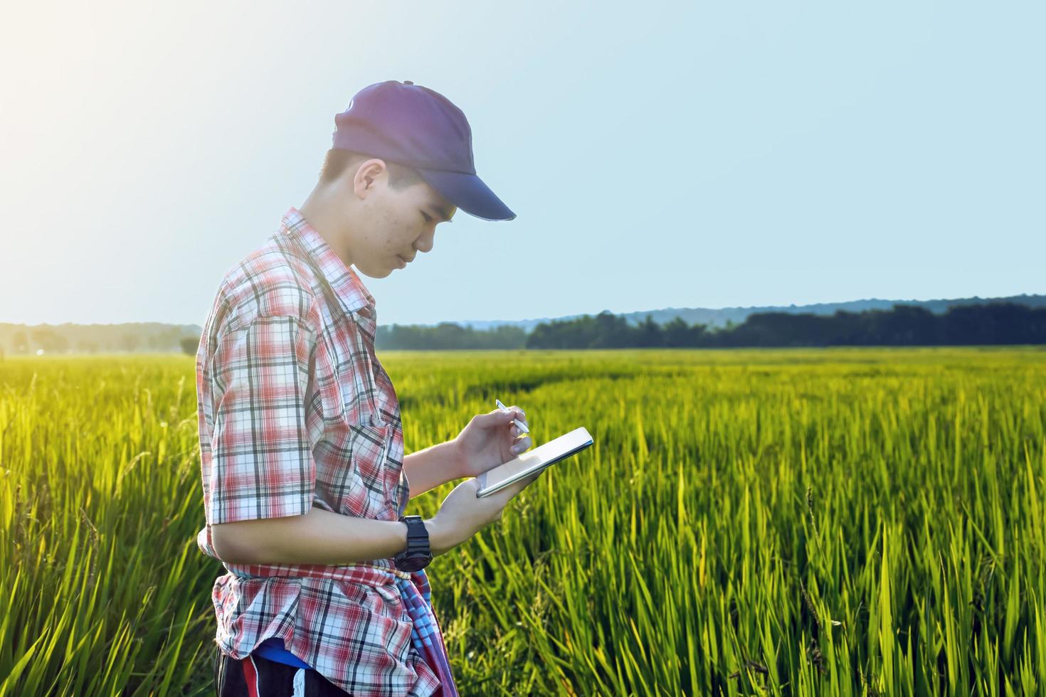 jovem adolescente asiático em camisa xadrez, usa boné e segurando o tablet nas mãos, de pé e usando seu tablet para pesquisar informações sobre o cultivo de arroz e fazer trabalhos de projeto escolar no arrozal. foto