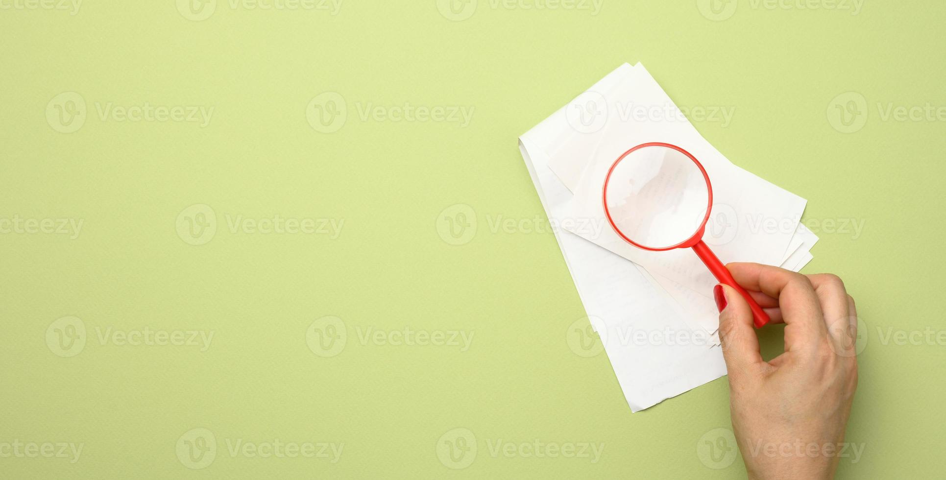 uma pilha de recibos de vendas em papel e a mão de uma mulher com uma lupa de plástico vermelha sobre um fundo verde. o conceito de controle de despesas e receitas foto