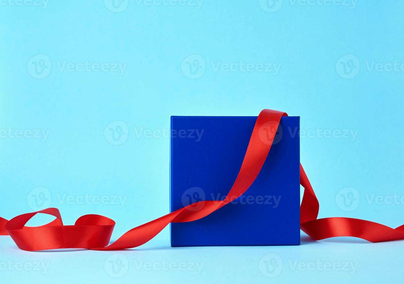 caixa de papelão quadrada azul para um presente e fita vermelha de seda torcida em um fundo azul foto