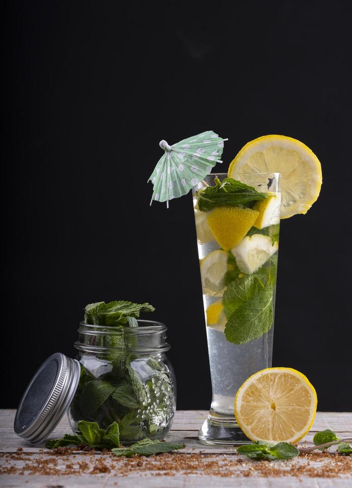 Coquetel de suco de limão com gelo e hortelã, acompanhado de jarra de vidro com hortelã sobre base de madeira. foto