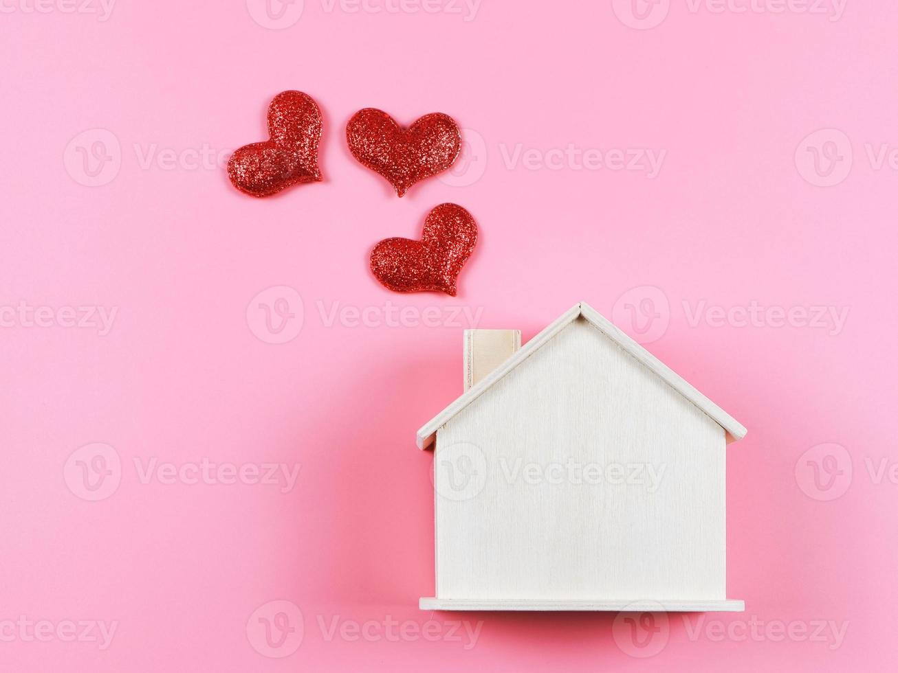 configuração plana da casa modelo de madeira com corações de glitter vermelho no fundo rosa. casa dos sonhos, lar do amor, relacionamento forte, dia dos namorados. foto