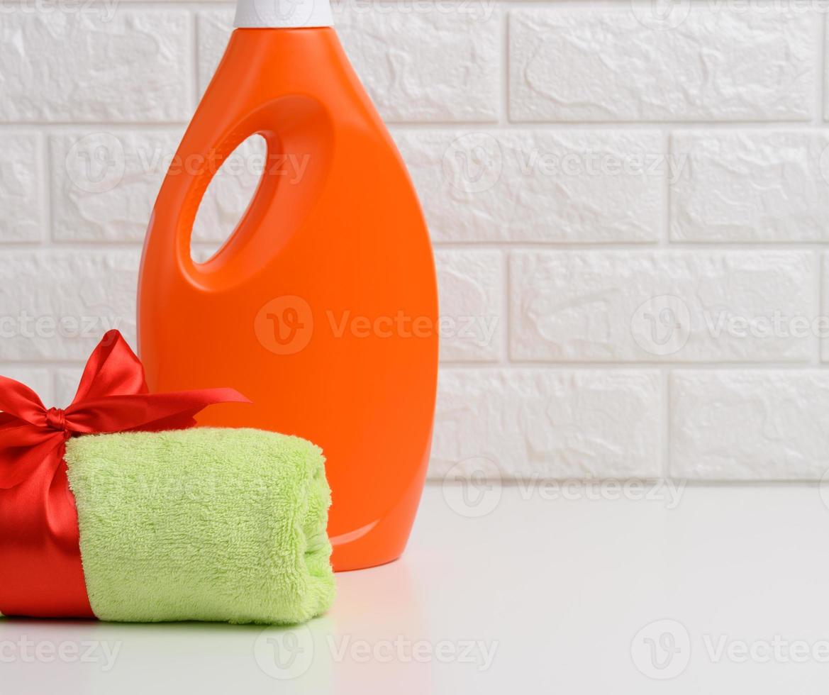 uma toalha verde felpuda enrolada amarrada com uma fita de seda vermelha e uma garrafa plástica laranja de detergente líquido em uma prateleira branca no banheiro foto