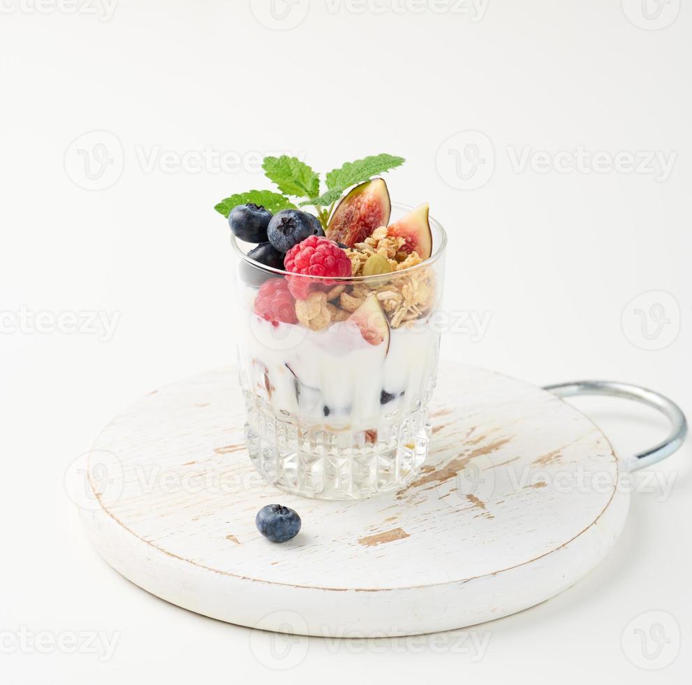 vidro transparente com granola derramada com iogurte, em cima de framboesas maduras, mirtilos e figos em uma mesa branca. café da manhã saudável foto