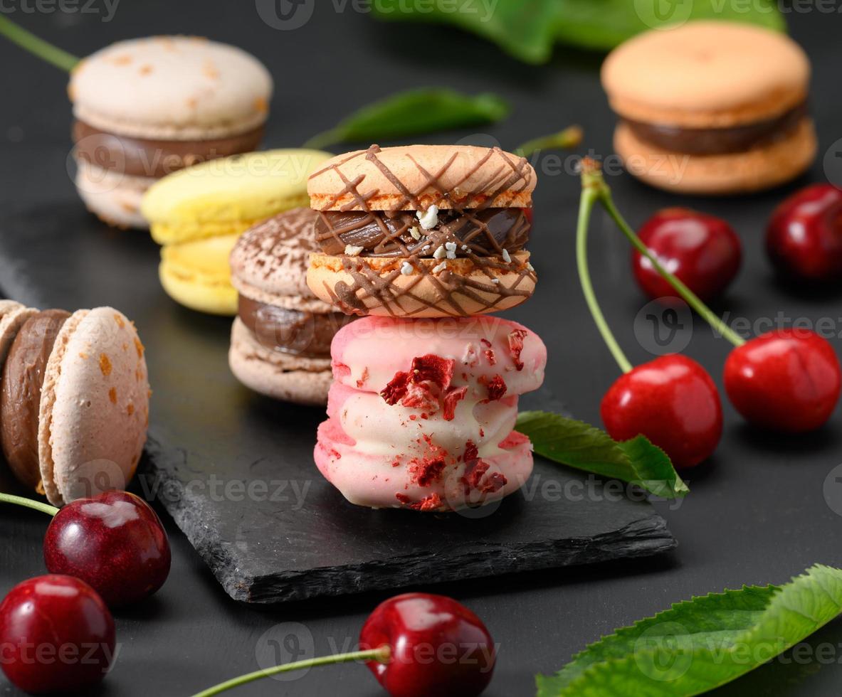 pilha de macarons multicoloridos e cerejas vermelhas maduras em fundo preto de madeira foto