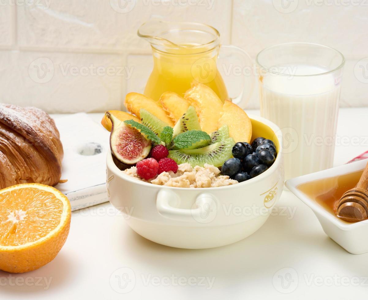 prato com aveia e frutas, meia laranja madura e suco espremido na hora em uma jarra de vidro transparente, mel em uma tigela sobre uma mesa branca. café da manhã saudável foto