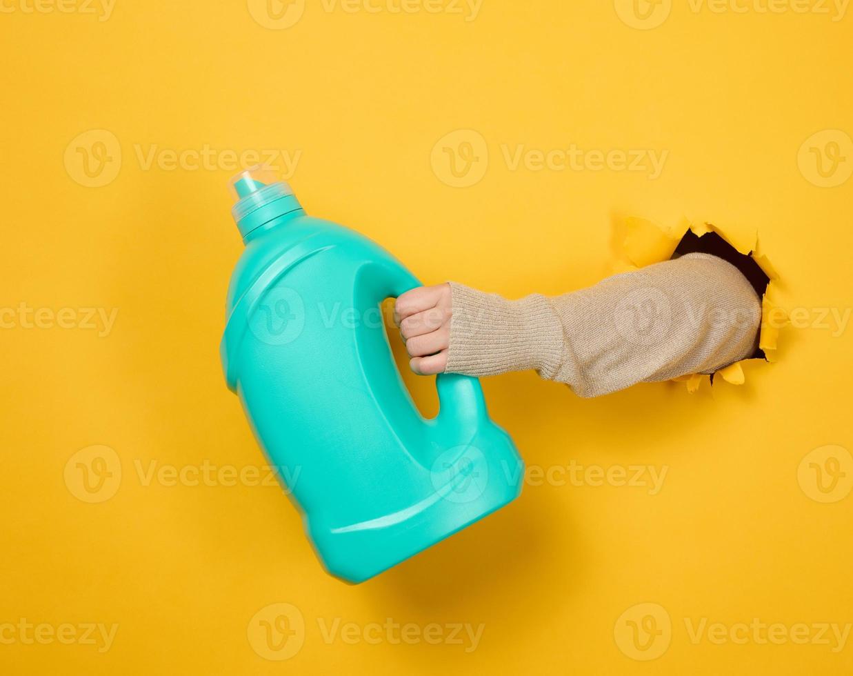 garrafa de plástico azul com detergente líquido em uma mão feminina em um fundo amarelo. uma parte do corpo sai de um buraco rasgado no fundo foto