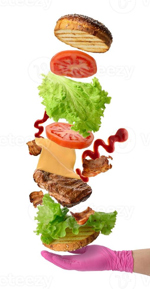 levitando ingredientes cheeseburger pão de gergelim, bife frito, salada verde e fatias de tomate. sanduíche sobre a mão na luva rosa foto