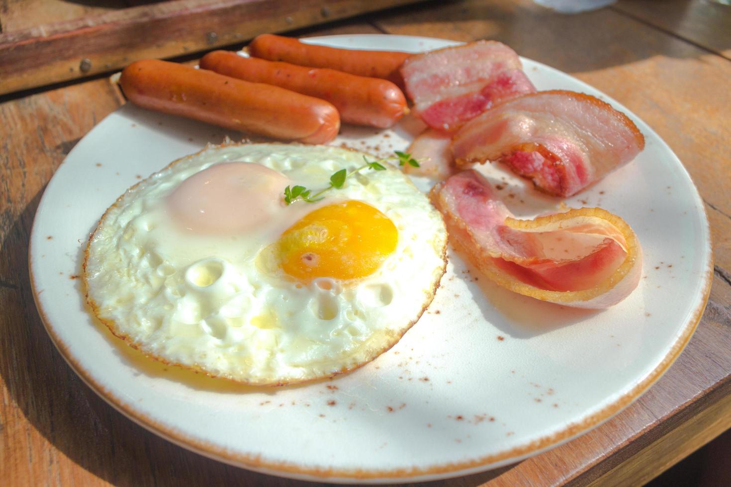 bacon, ovos fritos e salsicha no prato branco e mesa de madeira foto