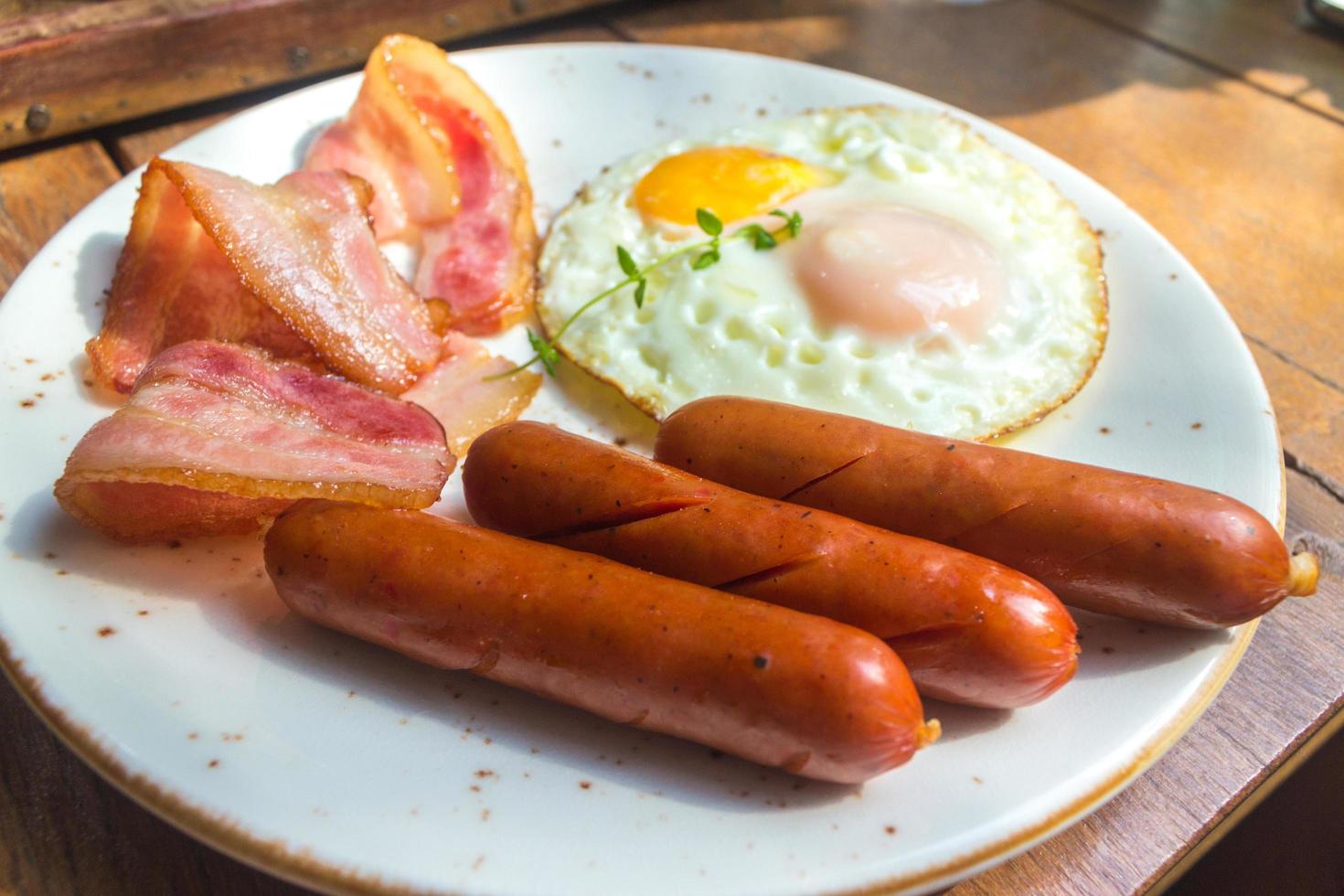 bacon, ovos fritos e salsicha no prato branco e mesa de madeira foto