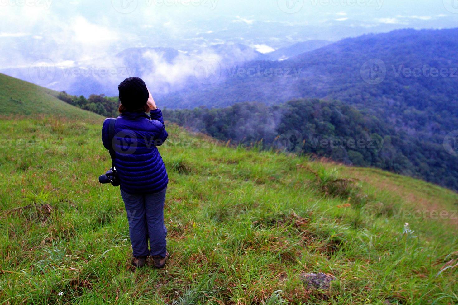 turista ou caminhadas no suéter azul com chapéu preto e segurando a câmera sozinho no campo de grama verde com ar fresco e névoa no fundo da montanha com espaço de cópia, vista de paisagem de beleza e viagens foto