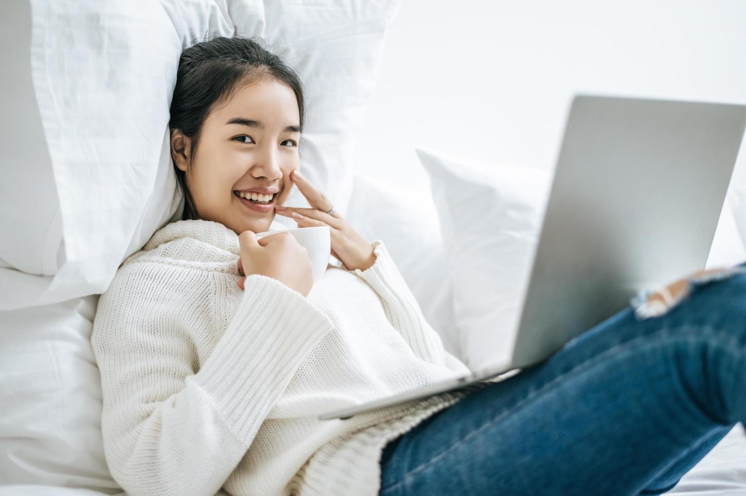 jovem brincando no laptop e segurando uma xícara de café na cama foto