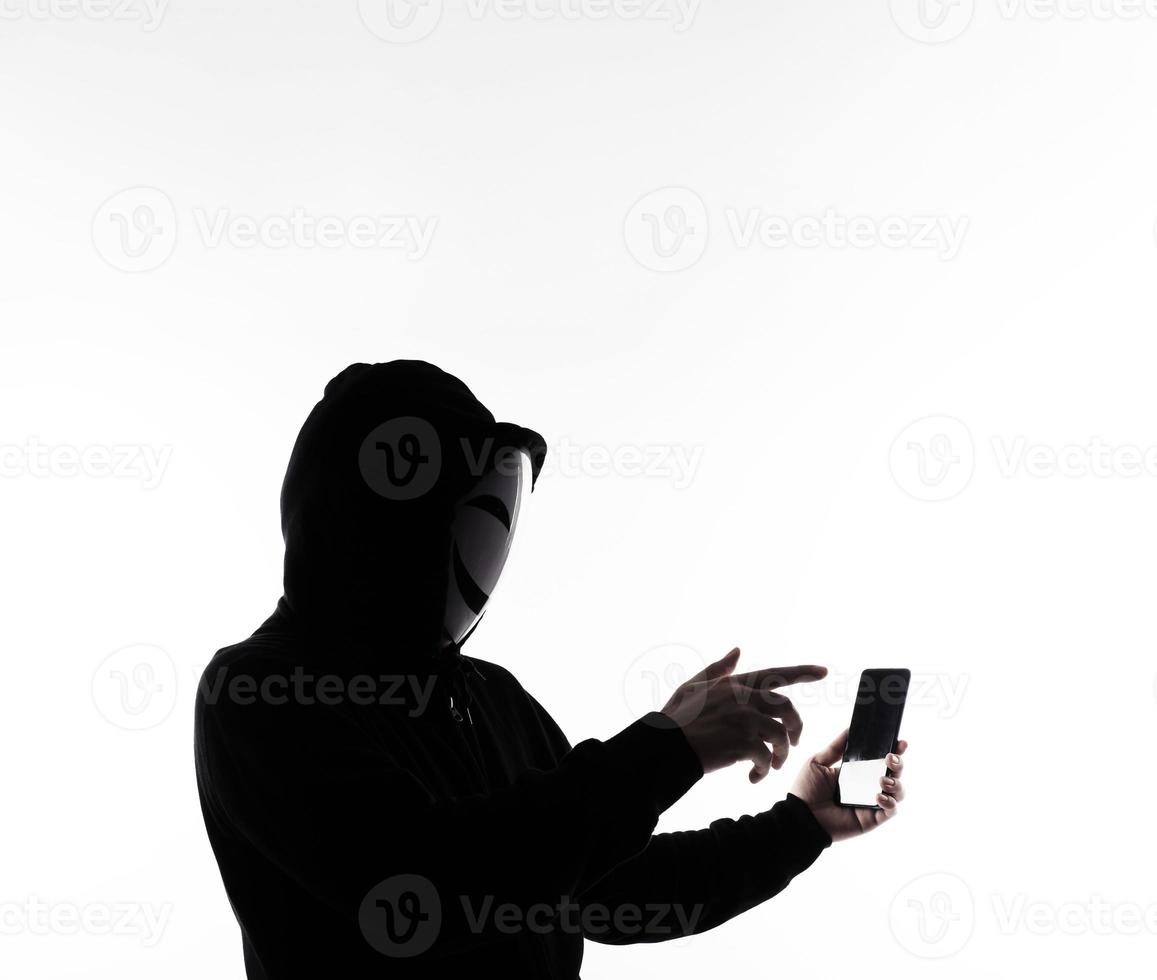 hacker anônimo e máscara facial com smartphone na mão. homem de camisa de capuz preto segurando e usando o celular em fundo branco. represente o hacking de dados de crimes cibernéticos ou o roubo do conceito de dados pessoais foto