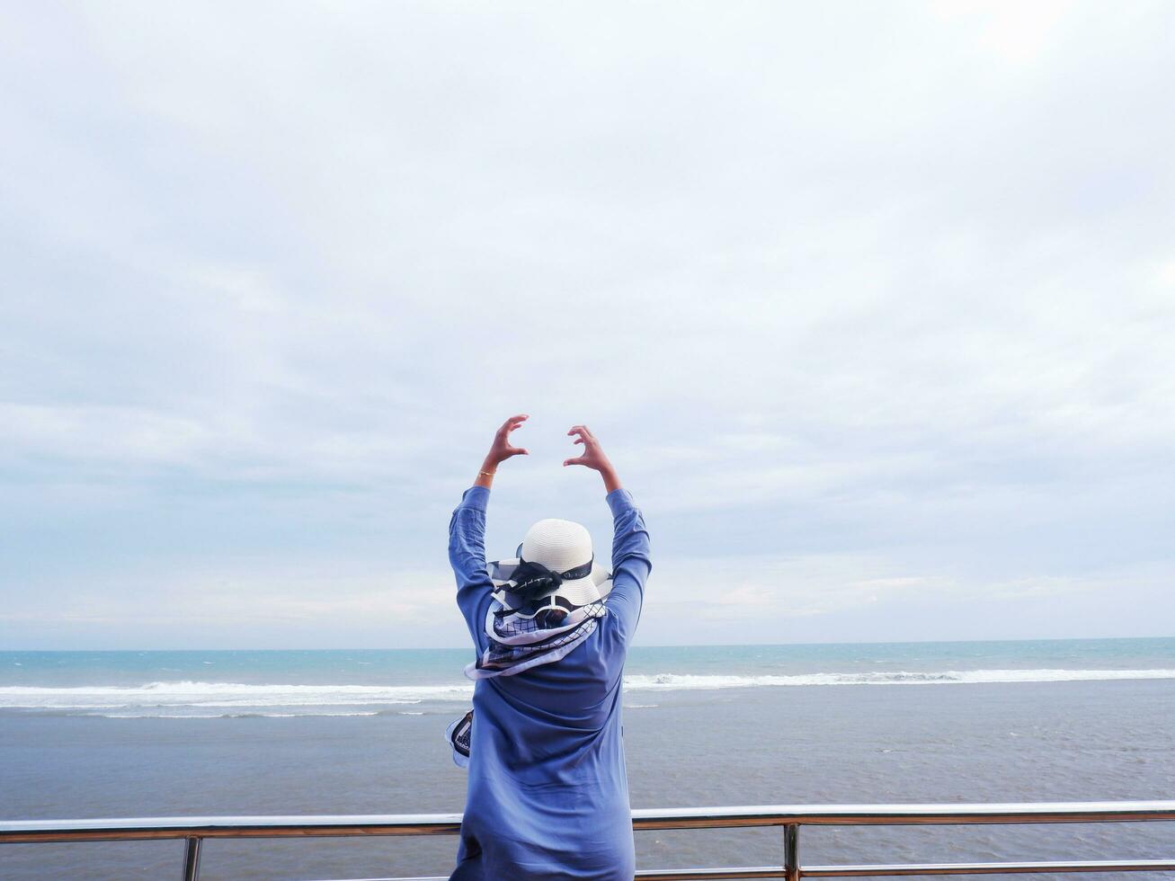costas da mulher de chapéu na praia tropical que olhava o céu e o mar enquanto suas mãos formavam amor na ponte. vista para praia foto