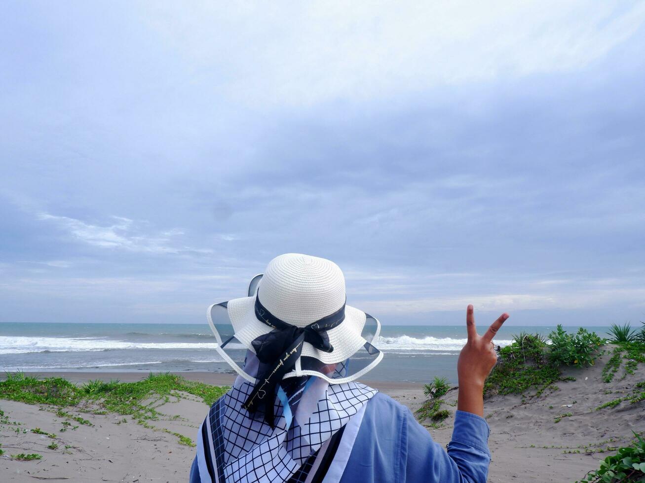 costas da mulher de chapéu na praia tropical que está olhando para o céu e o mar, mostrando o gesto de paz. vertical. fundo do mar. areia da praia foto