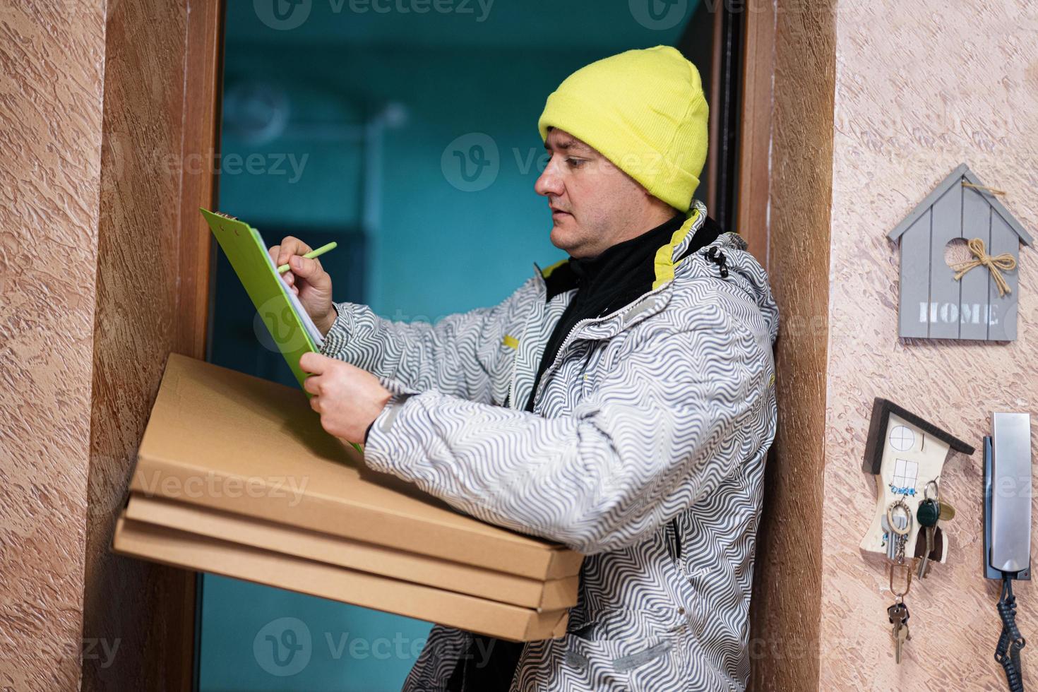 entregador com caixas de papelão de pizza. correio com chapéu de cor verde segurando uma prancheta em pé contra a porta da casa residencial foto
