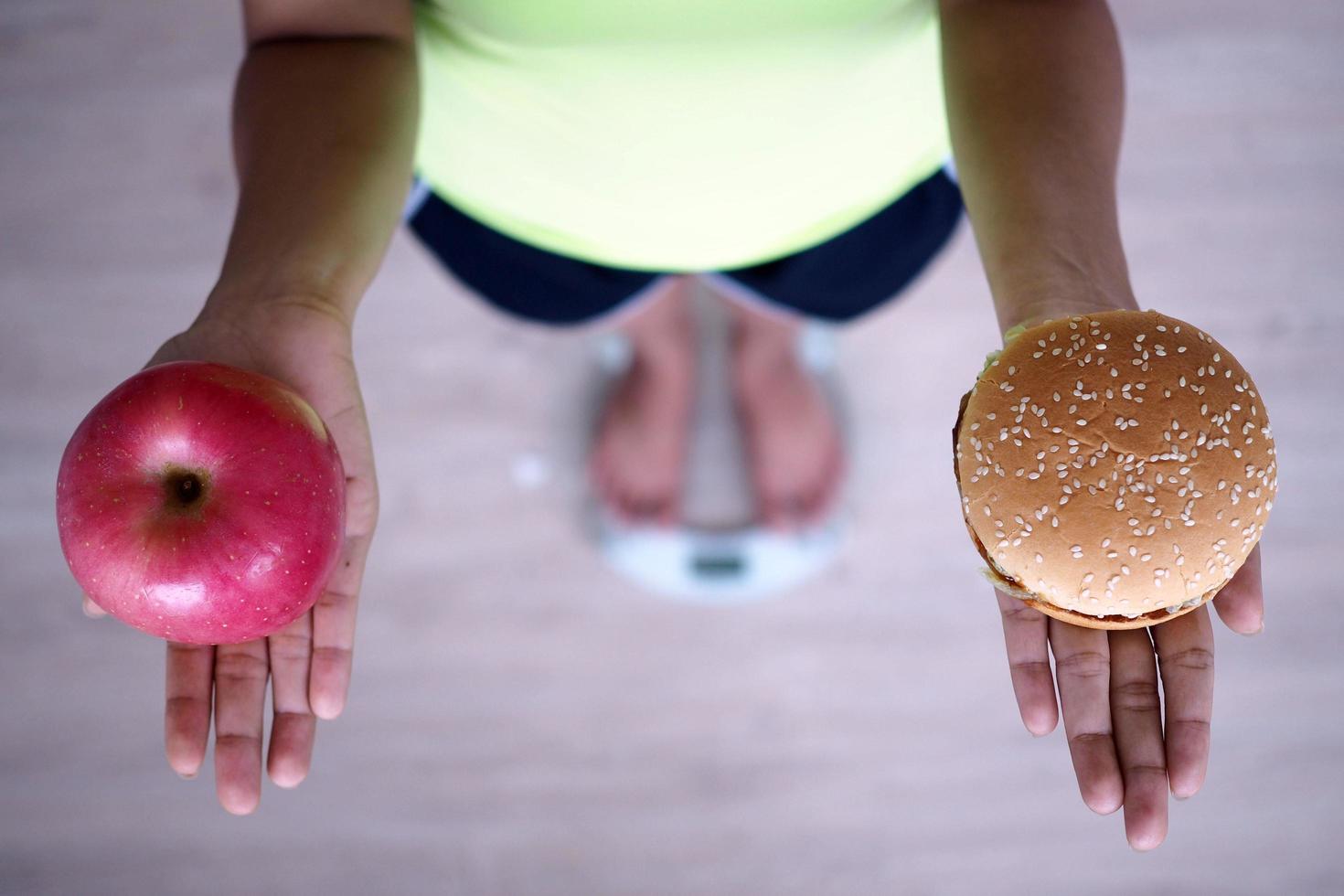 as mulheres estão pesando em balanças, segurando maçãs e hambúrgueres. a decisão de escolher junk food que não faz bem à saúde e frutas ricas em vitamina c fazem bem ao corpo. conceito de dieta foto