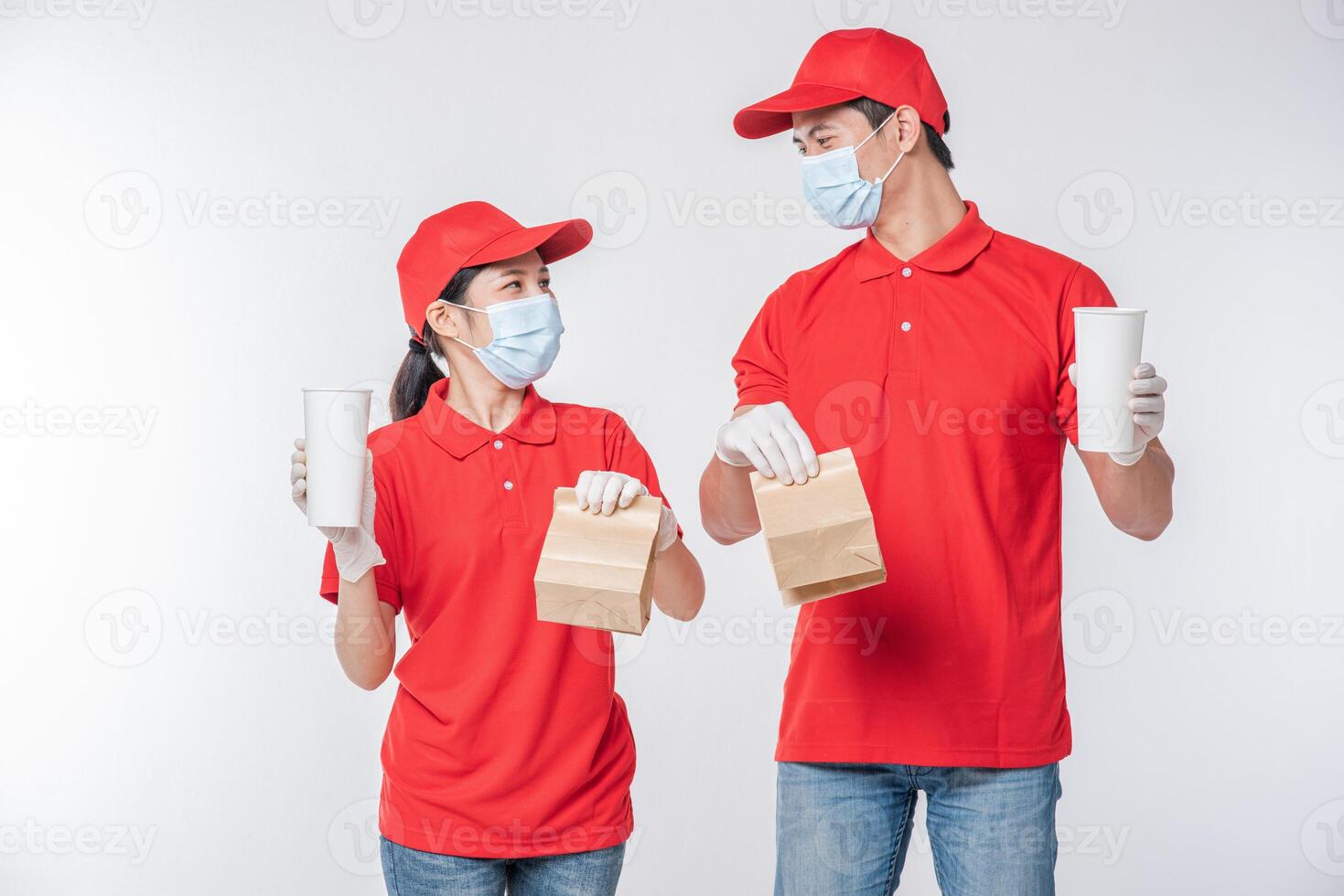 imagem de um jovem entregador feliz com gorro vermelho camiseta em branco uniforme máscara facial luvas em pé com pacote de papel artesanal marrom vazio isolado no estúdio de fundo cinza claro foto