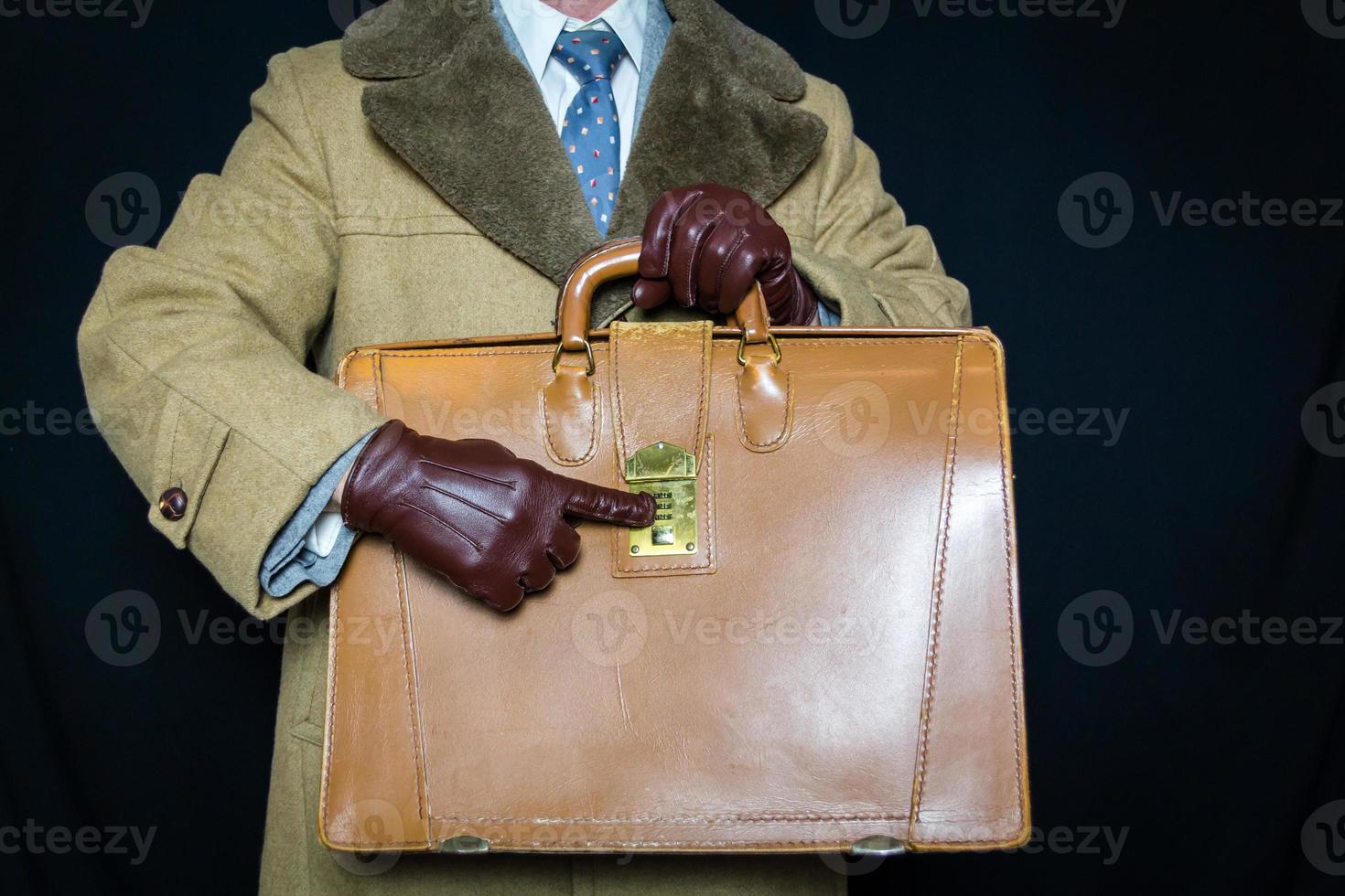 retrato de homem de casaco de pele marrom e luvas de couro apontando para combinação na maleta em fundo preto. conceito de agente secreto espião herói filme noir. roubo de identidade. foto