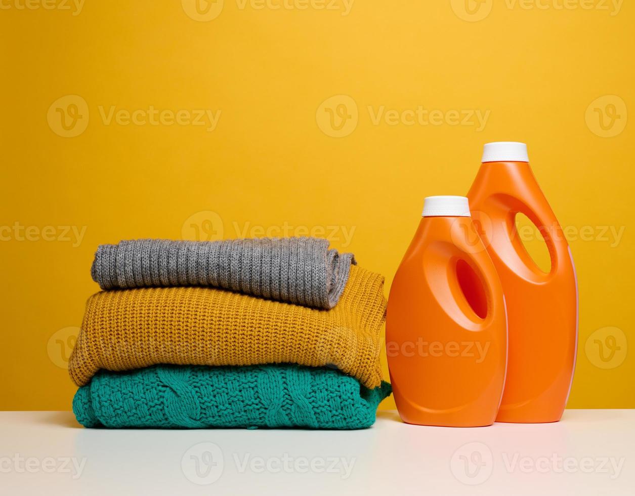 pilha do lavado guardada roupas e plástico laranja ampla garrafas com líquido detergente ficar de pé em uma branco mesa, amarelo fundo foto