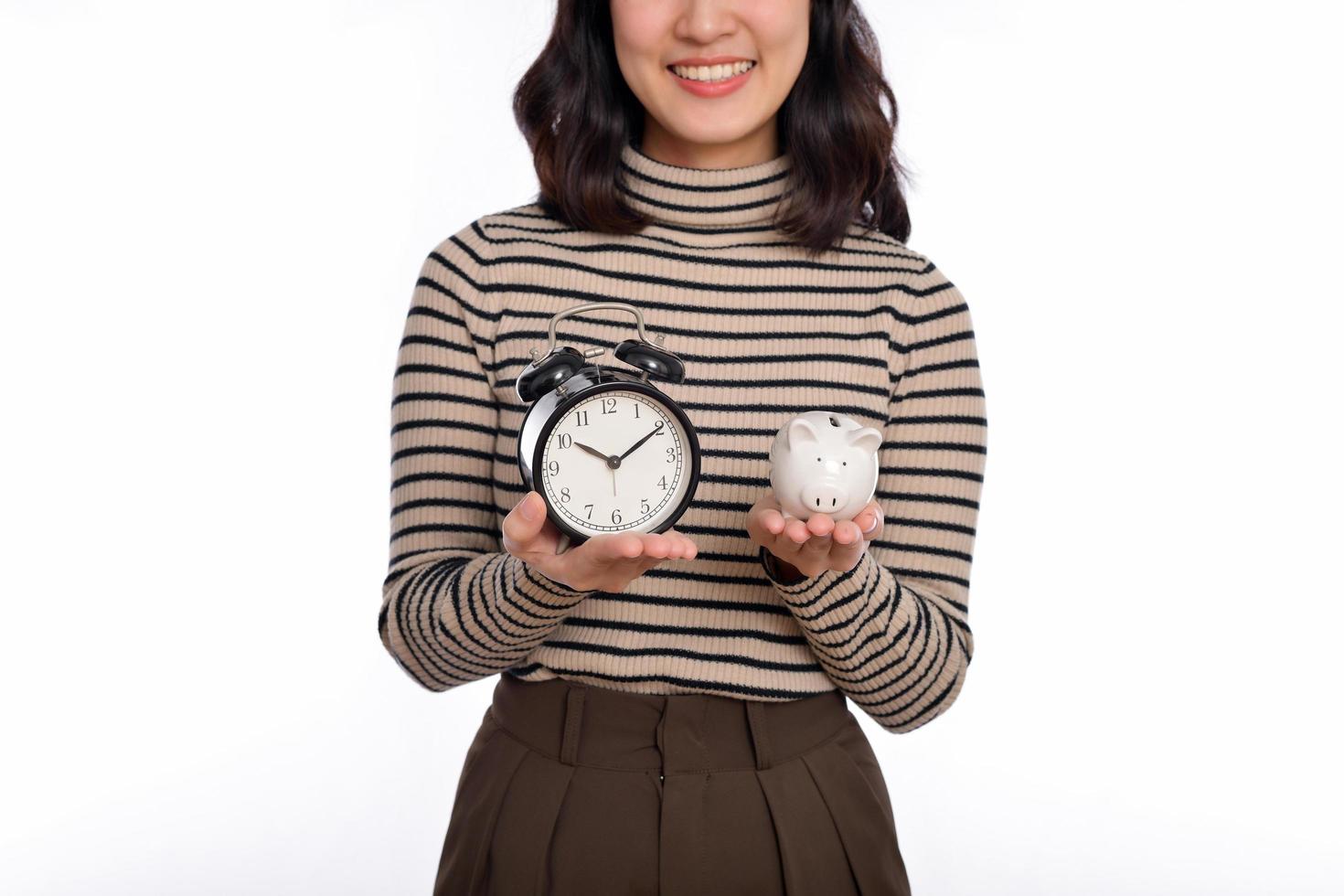 feliz ásia mulher com suéter camisa segurando alarme relógio e porquinho banco isolado em branco fundo foto