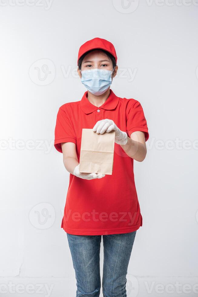 imagem de um jovem entregador feliz com gorro vermelho camiseta em branco uniforme máscara facial luvas em pé com pacote de papel artesanal marrom vazio isolado no estúdio de fundo cinza claro foto