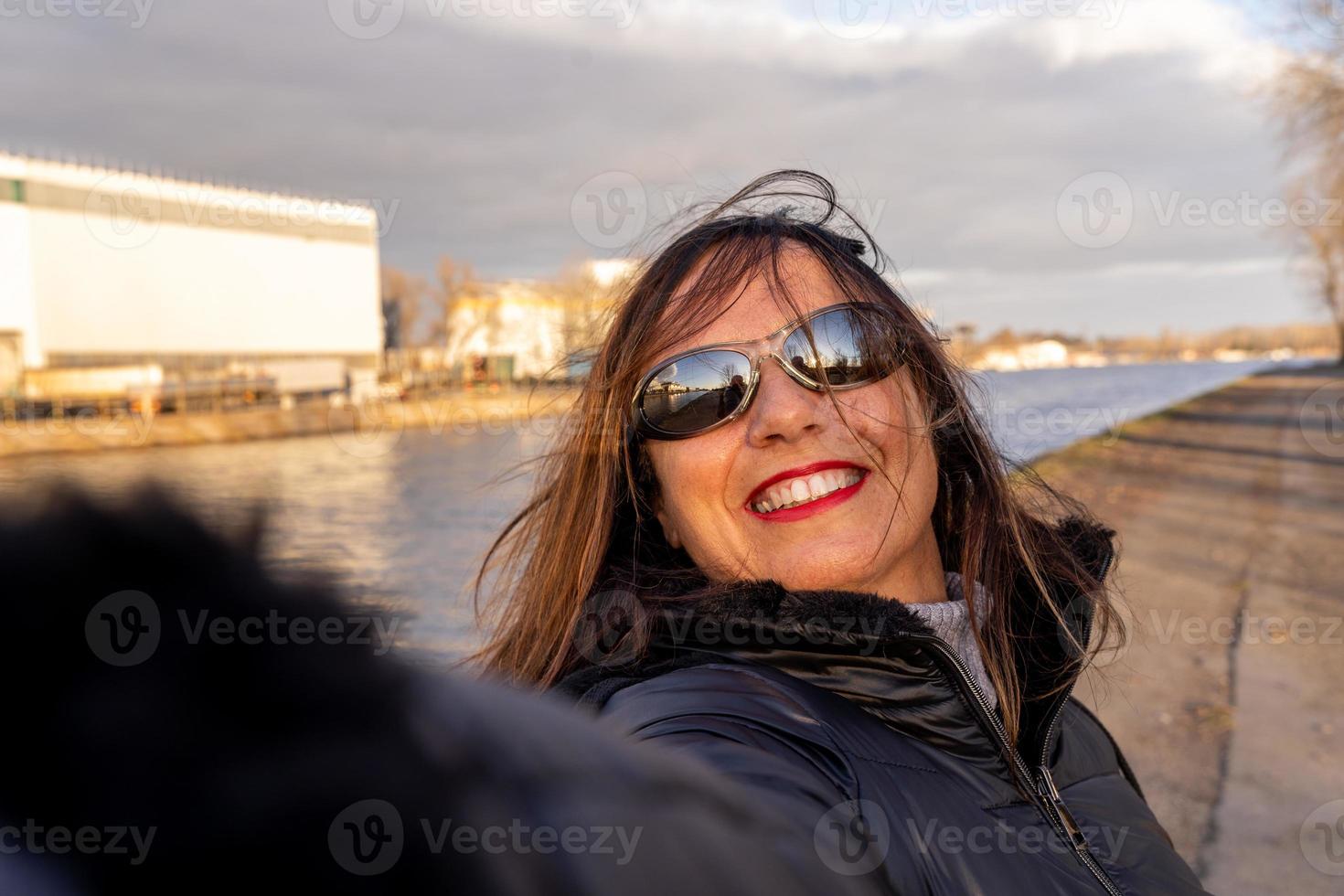 meio envelhecido mulher vestindo inverno roupas levando uma selfie de uma rio foto