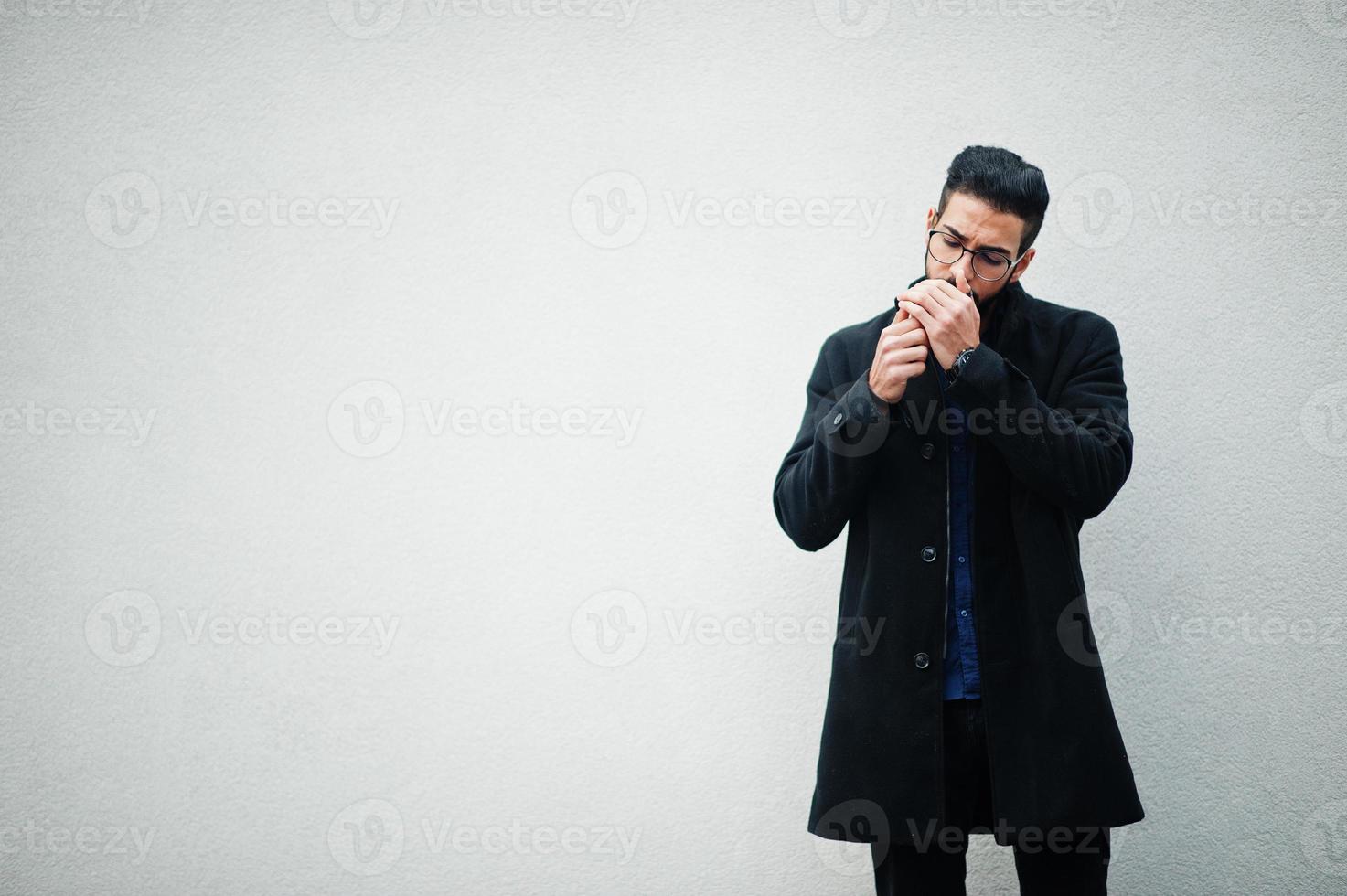 empresário do Oriente Médio usa casaco preto e camisa azul, óculos em pé contra a parede branca e fumando cigarro. foto