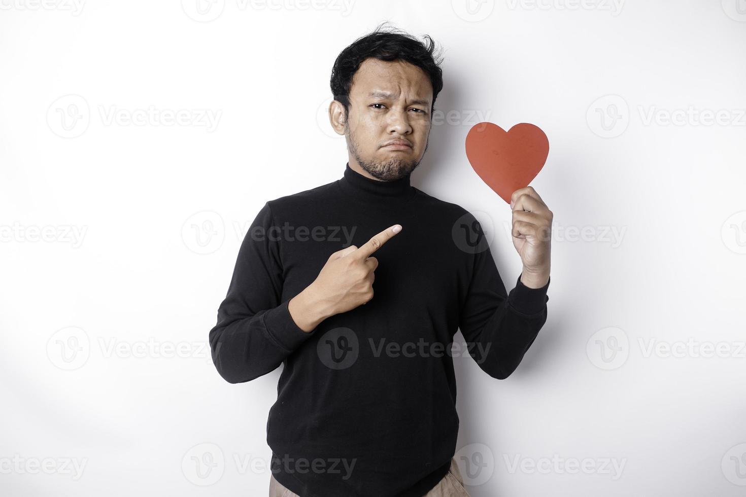 uma retrato do a ásia homem vestindo uma Preto camisa parece tão confuso enquanto segurando uma vermelho em forma de coração papel, isolado de uma branco fundo foto