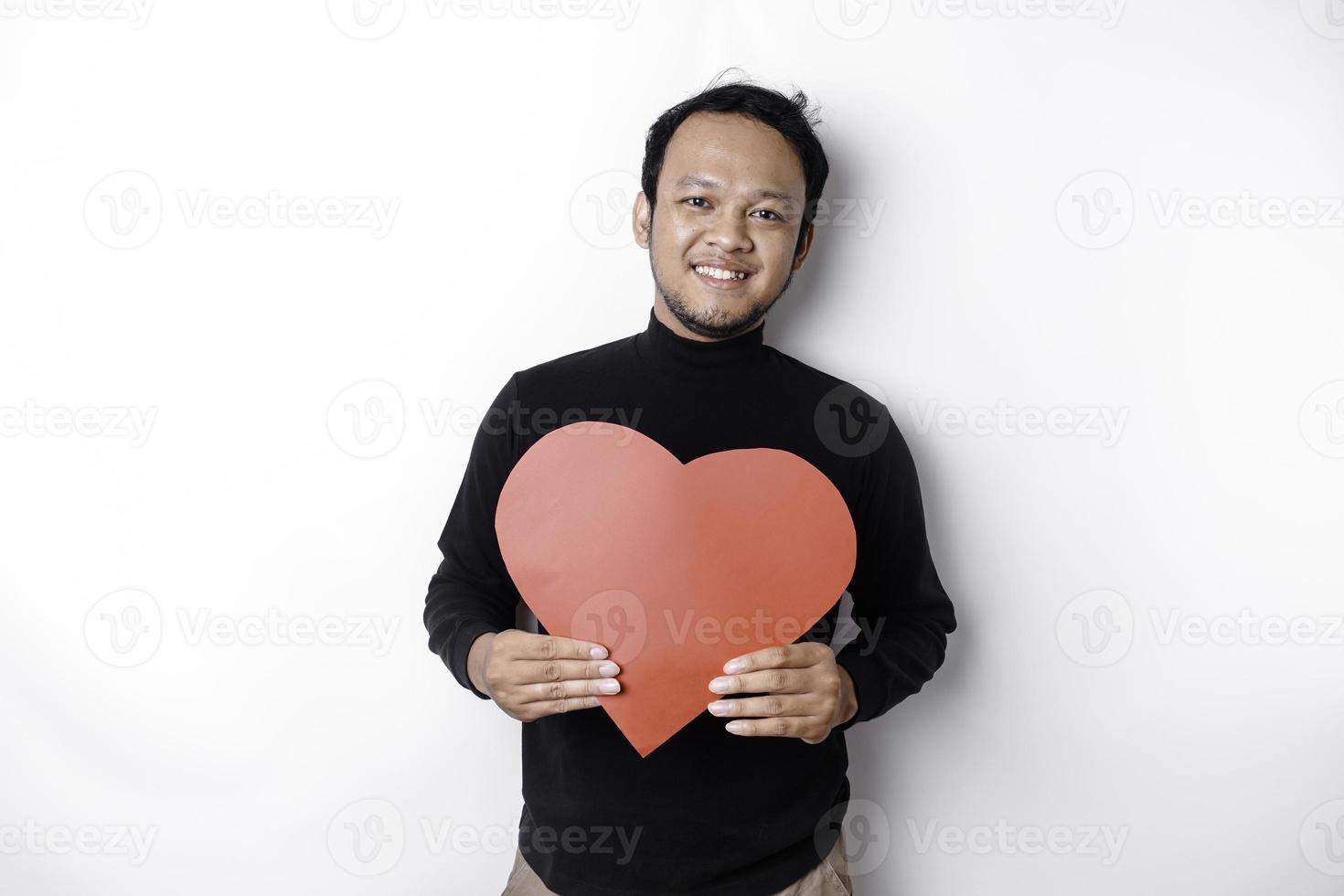 uma retrato do uma feliz ásia homem vestindo uma Preto camisa, segurando uma vermelho em forma de coração papel isolado de branco fundo foto