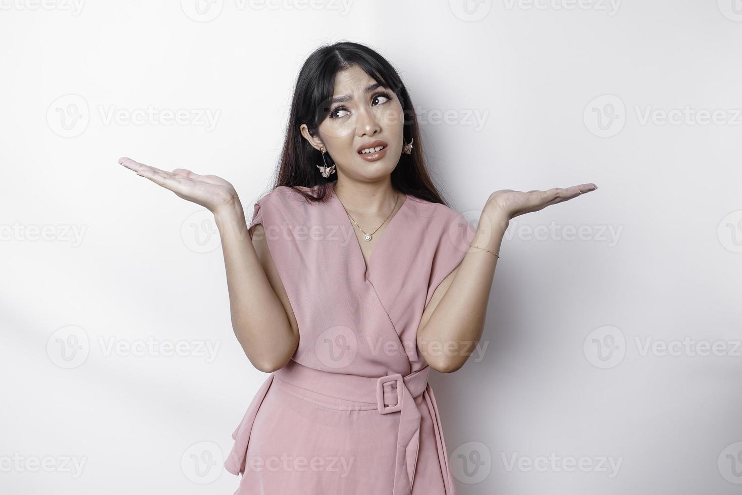 uma pensativo jovem ásia mulher é vestindo Rosa blusa segurando dela telefone e parece confuso, isolado de branco fundo foto