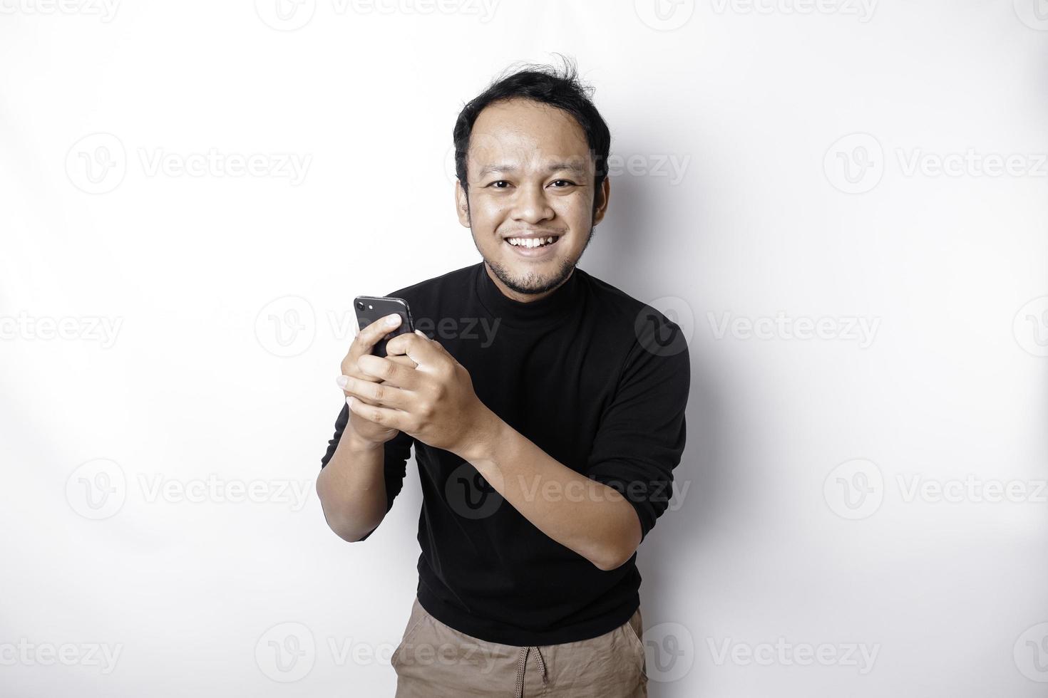 animado ásia homem vestindo Preto camisa sorridente enquanto segurando dele telefone, isolado de branco fundo foto