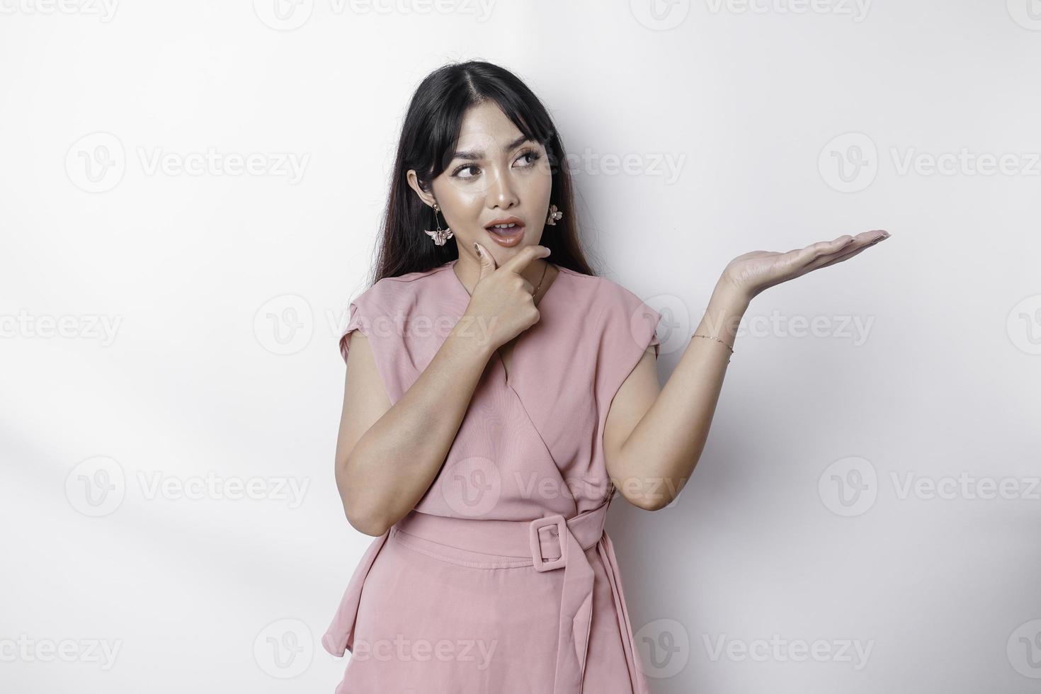 uma pensativo jovem mulher vestido dentro Rosa e segurando dela queixo enquanto apontando cópia de espaço ao lado dela, isolado de branco fundo foto