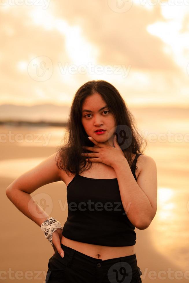 a ásia mulher com Preto roupas e grandes Preto cabelo em pé em a de praia areia com uma plano expressão enquanto em período de férias foto