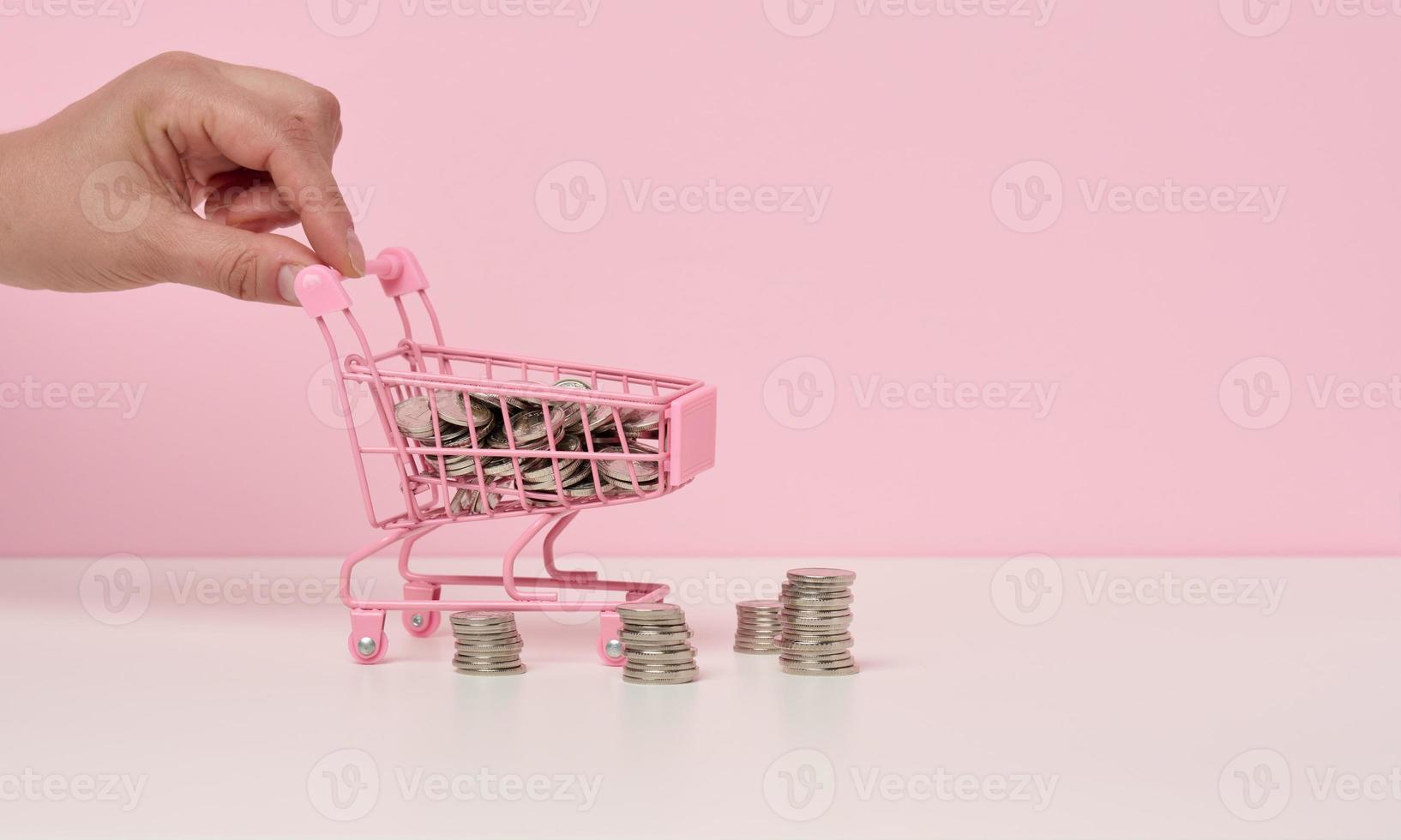 fêmea mão empurra uma miniatura metal compras carrinho e uma pilha do moedas em uma branco mesa. a conceito do descontos e vendas, despesas poupança. conectados comércio foto