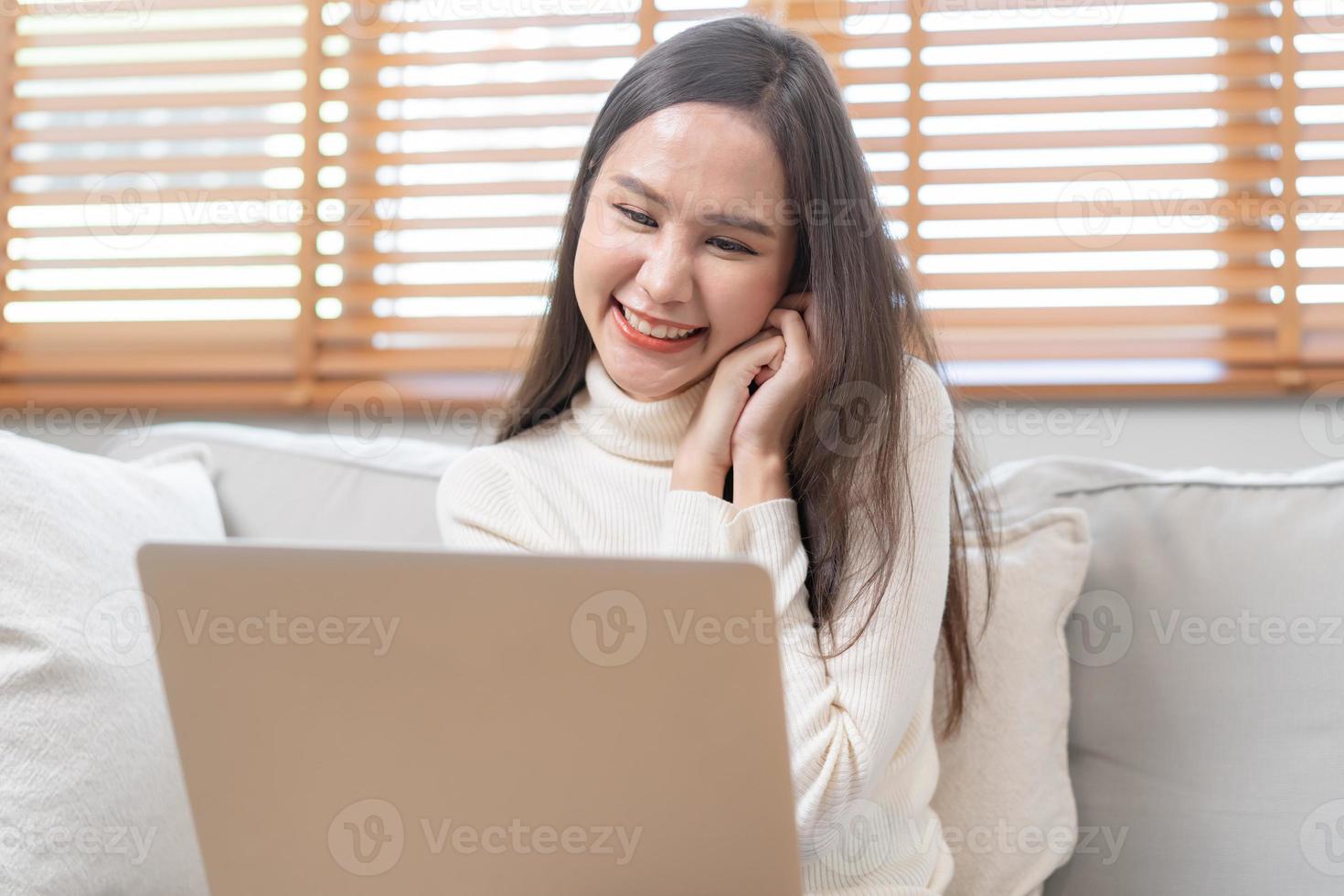 feliz alegre, satisfeito ásia jovem trabalhador autonomo mulher, menina sentado em sofá ou sofá, usando computador portátil computador dentro vivo quarto às lar, olhando e sorridente às tela, empreendedor desfrutando com trabalhar, wfh foto