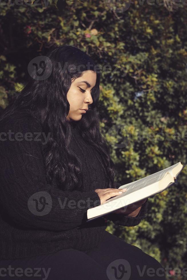 jovem mulher, sentado lendo uma livro dentro a jardim foto