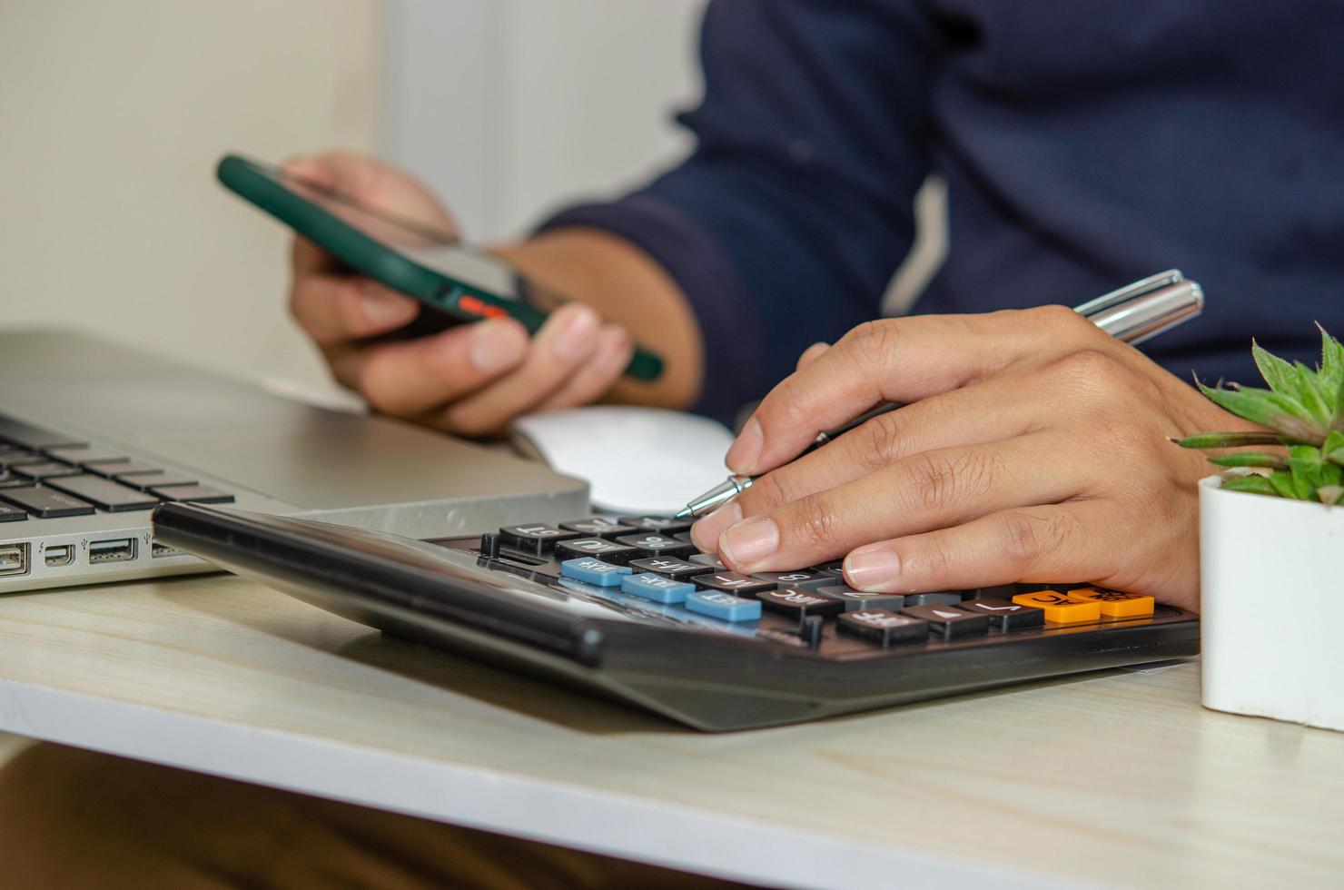 um empresário usando uma calculadora, segurando uma caneta e um telefone celular em uma mesa com um laptop trabalhando em casa foto