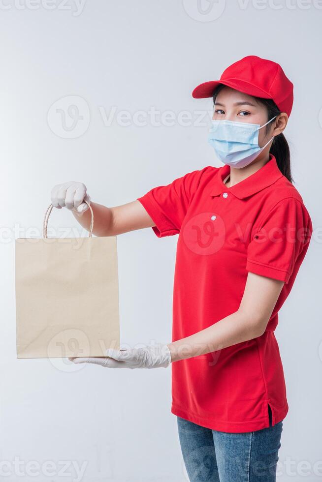 imagem de um jovem entregador feliz com gorro vermelho camiseta em branco uniforme máscara facial luvas em pé com pacote de papel artesanal marrom vazio isolado no estúdio de fundo cinza claro foto