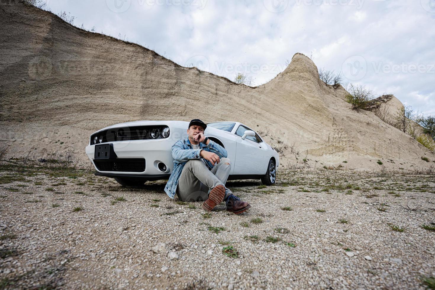 homem bonito em jaqueta jeans e boné está sentado perto de seu muscle car branco na carreira. foto