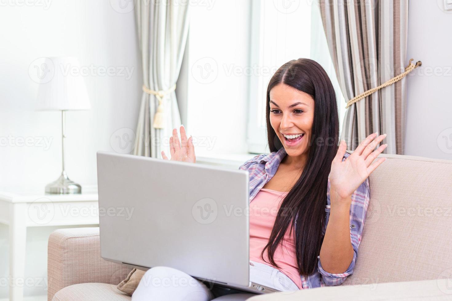 atraente jovem feliz sentada no sofá usando o laptop, conversando por vídeo, acenando, conversando com sua amiga sobre coronavírus e ficando em casa durante a videochamada online. trabalhando em casa foto