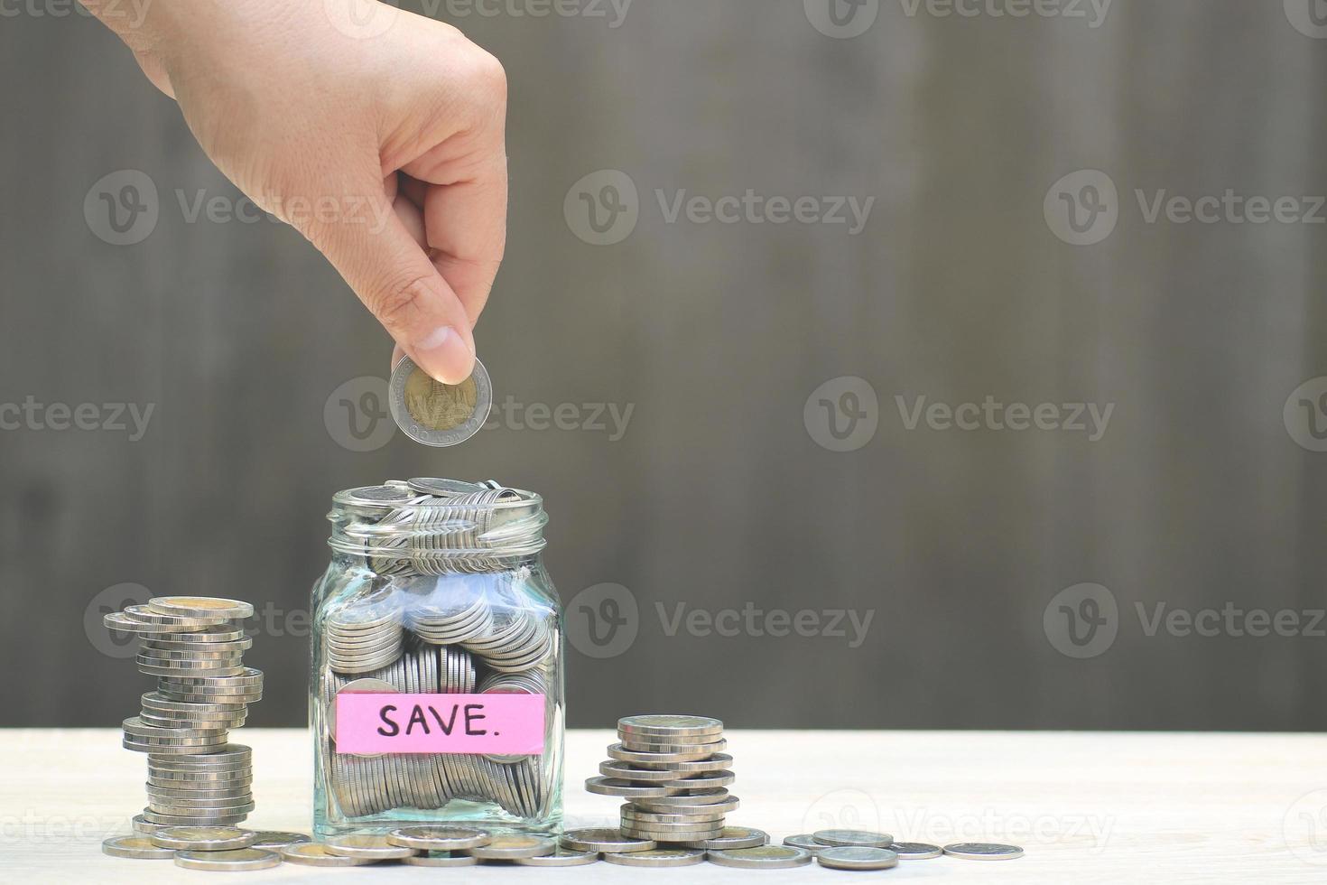 mão colocando a moeda em um frasco rotulado salvar ao lado de pilhas de moedas foto