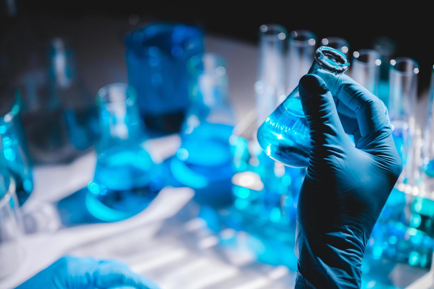 mão na luva azul segurando um frasco de líquido azul com frascos e frascos de líquido azul no fundo foto