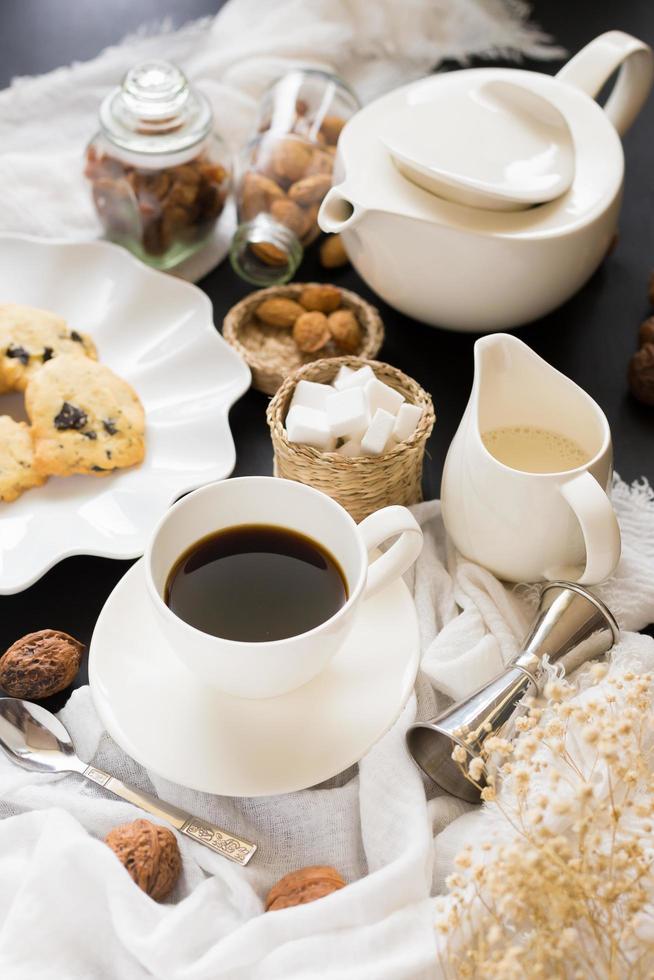 xícara de café, pires e servidor de creme ao lado de bolos na toalha de mesa amarrotada foto